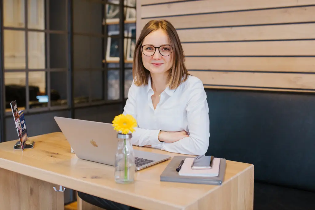 Sophie Gulich sitzt mit Brille und weißer Bluse lächelnd an einem Holztisch in einem Café, vor ihr ein aufgeklappter Laptop, ein Notizbuch mit Smartphone und eine Vase mit einer gelben Gerbera. finanzen-net-zero-erfahrungen