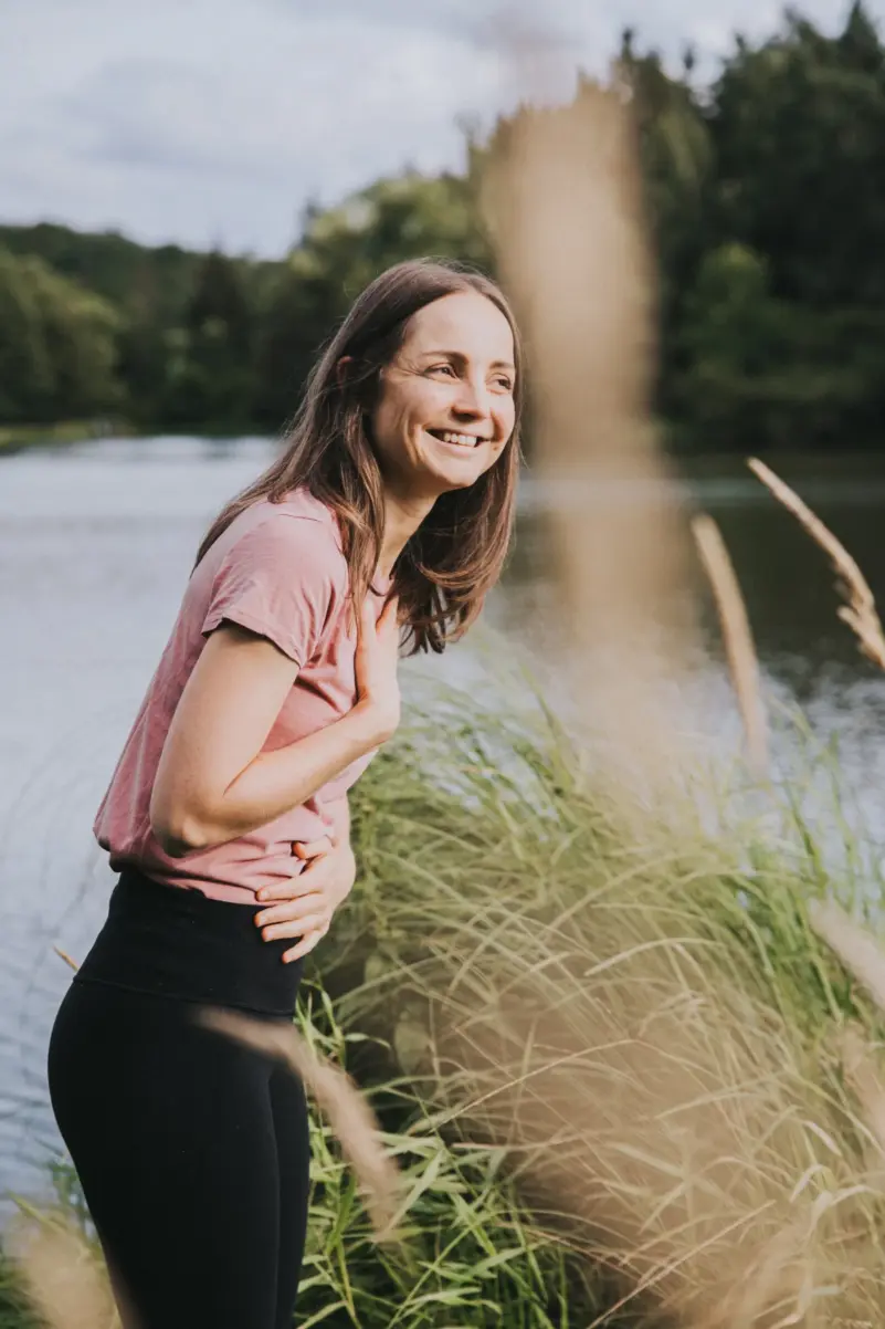 Selbstbestimmtes Leben: Sophie Gulich steht lachend am Seeufer im hohen Gras, eine Hand auf der Brust, eine auf dem Bauch.