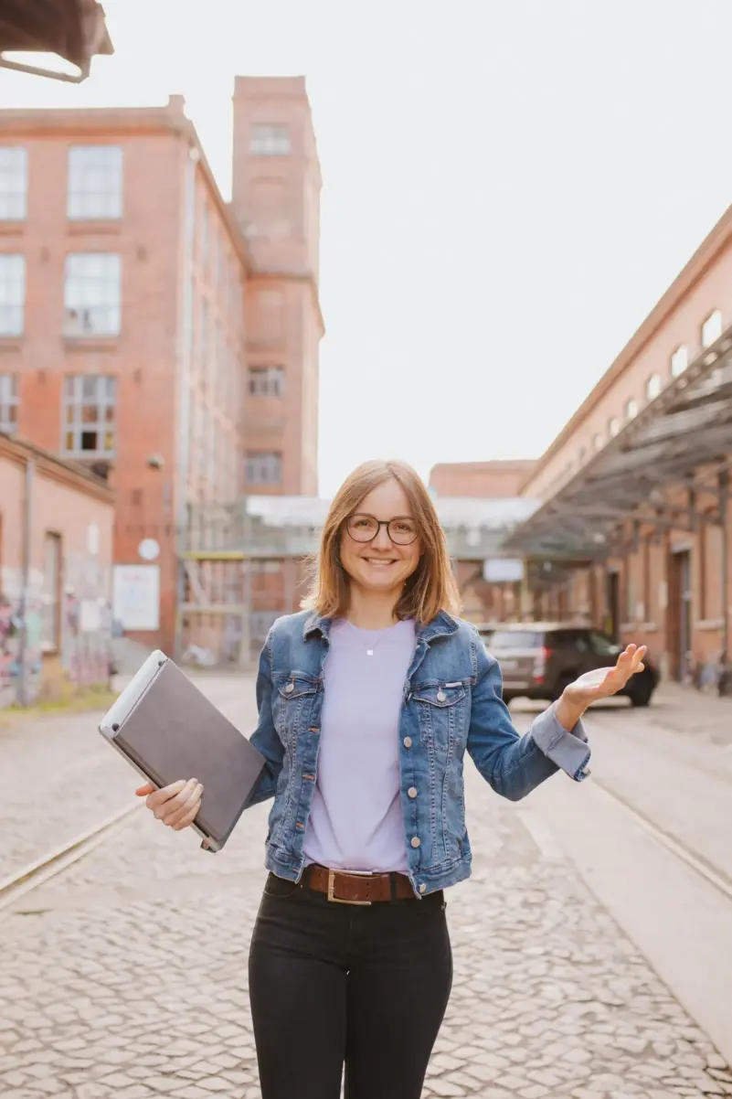 Selbstbestimmtes Leben: Sophie Gulich in Jeansjacke und Brille mit Laptop unter dem Arm, lächelnd vor einem alten Backstein-Fabrikgelände.