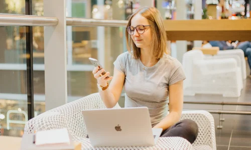 Bondora-Go-and-Grow-Erfahrung, Sophie Gulich sitzt mit Brille und grauem T-Shirt im Schneidersitz in einem weißen Geflechtsessel, schaut auf ihr Smartphone und hat einen aufgeklappten Laptop vor sich, im Hintergrund ein heller Innenraum mit Glasgeländer.