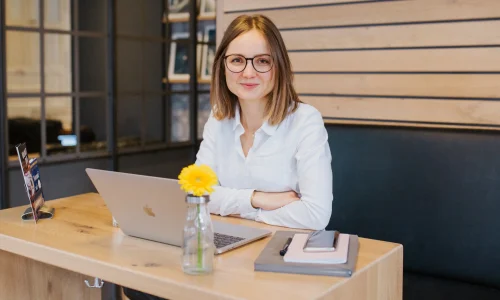 Sophie Gulich sitzt mit Brille und weißer Bluse lächelnd an einem Holztisch in einem Café, vor ihr ein aufgeklappter Laptop, ein Notizbuch mit Smartphone und eine Vase mit einer gelben Gerbera. finanzen-net-zero-erfahrungen
